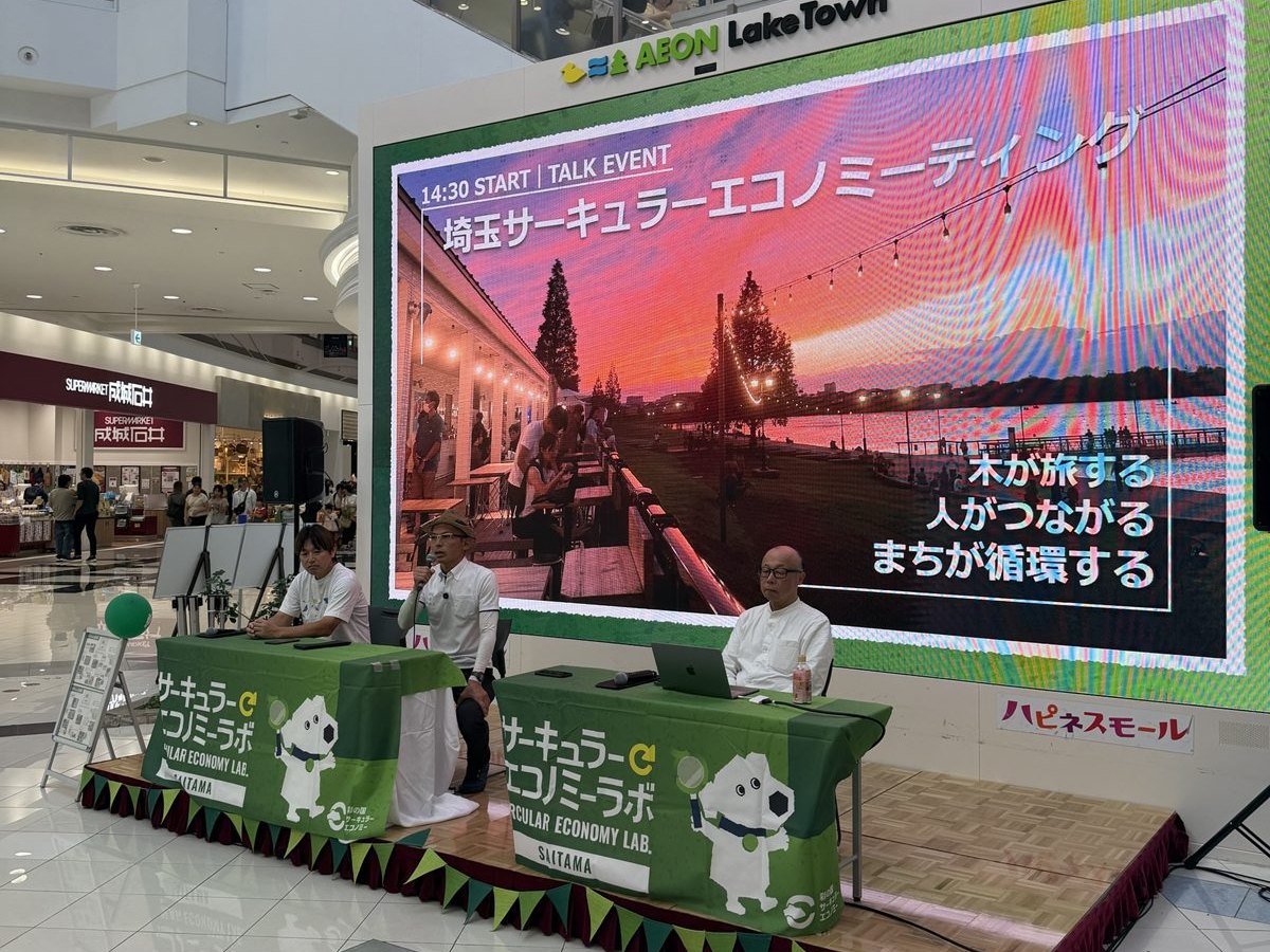 イオンレイクタウンのステージ。大きなスクリーンに「木が旅する 人がつながる まちが循環する」と映り、その前のテーブルに登壇者3人が並ぶ